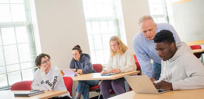 Professor Joseph D'Adamo helps a student in class, leaning down to look at the student's open laptop with him